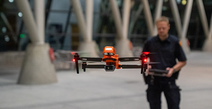 Officer in uniform holding the controls of a red drone flying toward the screen.
